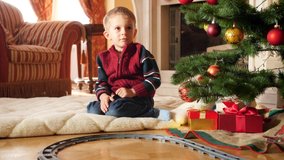 4k video of happy smiling little boy lying on floor at living room decorated for Christmas and playing with toy train and railway. Child receiving gifts and presents from Santa Claus on winter