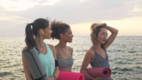 Group of cheerful young sporty multiethnic women friends in sportswear smiling and talking while walking with mats near the sea