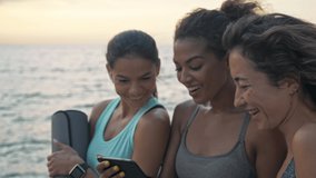 Group of smiling young sporty multiethnic women friends in sportswear talking and laughing while watching something on smartphone staying near the sea
