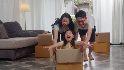 Happy Asian young family having fun laughing moving into new home. Japanese parents mother and father smiling helping excited girl riding in cardboard box. New property and relocation. Slow motion.