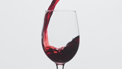 Red wine forms beautiful wave. Wine pouring in wine glass over white background. Close-up shot. Slow motion of pouring red wine from bottle into goblet.