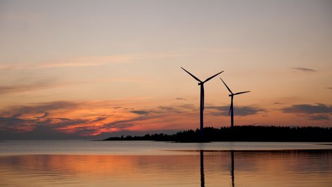 Two wind turbines, part of island based wind farm, spin at dusk. Silhouettes long shot at real wind speed, in Finland near Baltic sea shore.