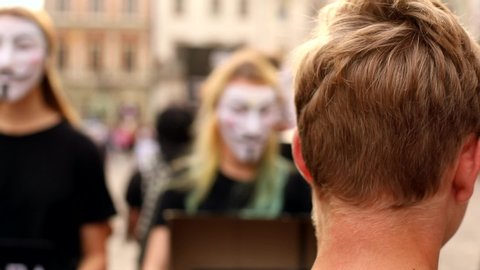 Lviv, Ukraine - September 6, 2019: Back view of young man standing in front of people with hidden faces by anonymous mask on sunny street. Smile White Hacker masks. Unique design of modern art object.