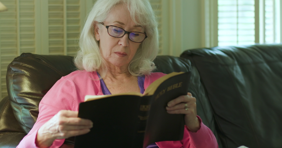 A middle aged Caucasian woman relaxing on a couch reading from a large traditional style Christian Bible stops and begins to pray.