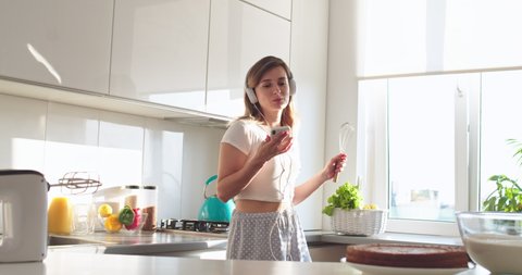 Caucasian cheerful young woman in the pajama and handphones listening to the music on the smartphone and dancing with mixer part in hand while preparing a breakfast in the morning in the kitchen.
