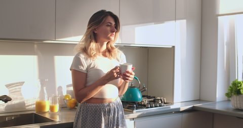 Pretty Caucasian young woman in pajama standing in the sunny kitchen in the morning, drinking hot coffe or tea and enjoying her time. Stay at home, quarantine, pandemic, coronavirus ,virus