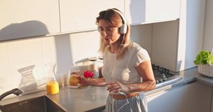 View from above on the young attarctive Caucasian woman in headphones dancing and listening to the music on the smartphone player while sipping orange juice in the kitchen.