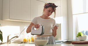 Caucasian attractive young woman housewife churning a cream with a mixer in a bowl and reading a recipe or watching video on the smartphone while preparing cooking in the kitchen.