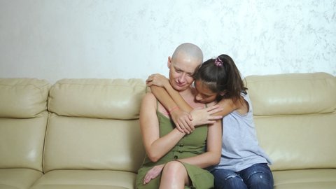 bald exhausted mother after oncology chemotherapy hugs her teenager daughter while sitting on the couch
