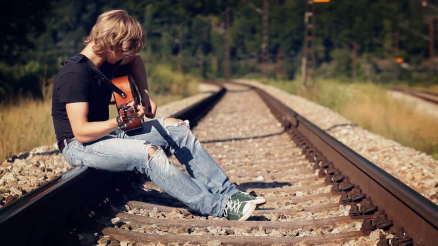 Long-haired guitarist wearing sunglasses and ripped jeans sitting on railroad playing his instrument