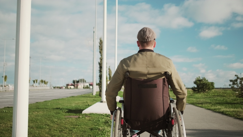 Adult disable man in invalid carriage is riding in park in sunny day, back view. He is enjoying good weather, after rehabilitation