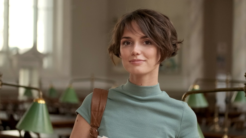 Portrait of pretty smiling female student joyfully looking in camera in library of university 