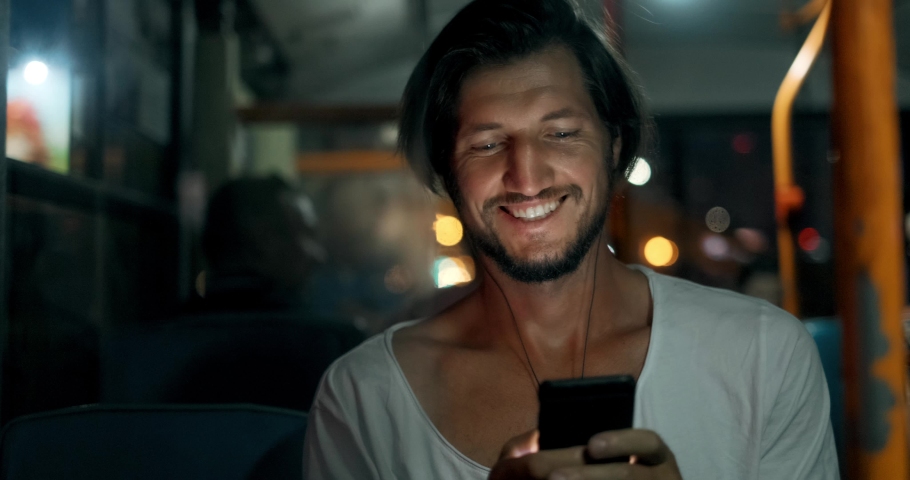 Young man holds smartphone and smiling while traveling by bus at night. Bearded male in headphones with phone, touches screen with fingers and looks out window