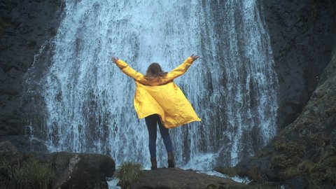 Young girl traveler hiker in a yellow raincoat walks to a waterfall in the highlands, raises his hands up, enjoys nature and life. Traveling in the mountains, adventure in trip. Lifestyle concept.