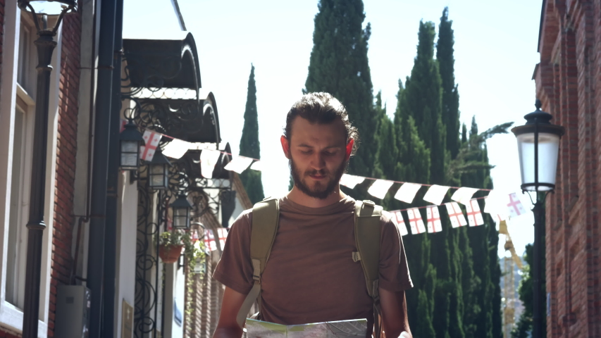 Young guy traveler is walking in a European city with a map in his hands. A tourist is looking for a way or lost. Sightseeing in the new city. Man traveler reading geographical atlas.