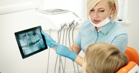 Professional female dentist showing preteen boy x-ray scan on tablet and explaining problem, modern technologies