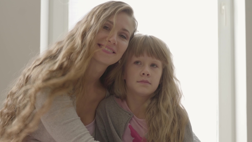 Smiling young woman looking at camera and hugging the sleepy little girl. Cucasian mother spending time with a cute blonde daughter.