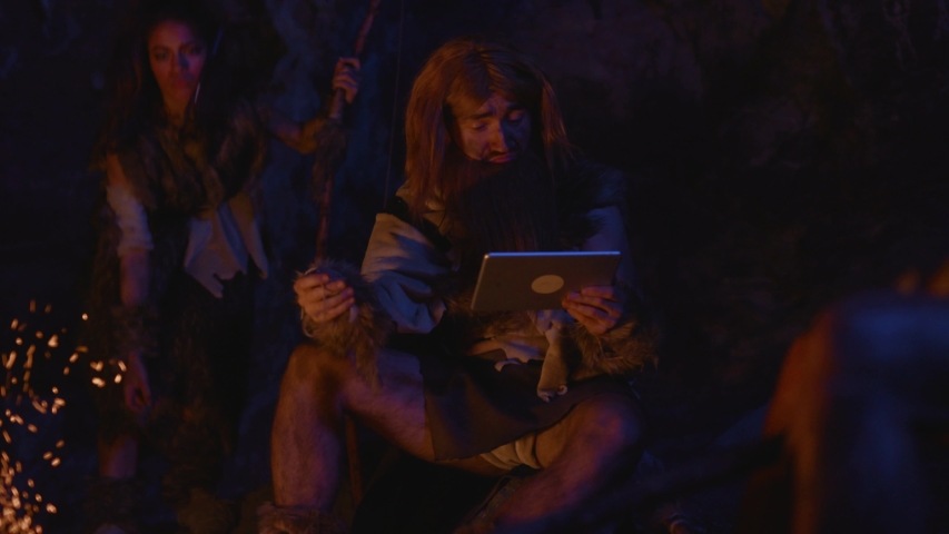 Cave people. Family of ancient wild people at bonfire outside cave dwelling. Excited fun tribe leader using tablet app like modern man. Homo sapiens.