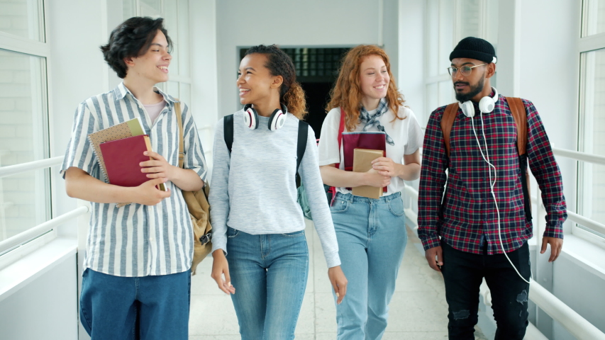 Cheerful students multi-racial group are talking walking with books in college hall showing thumbs-up laughing. Emotions, lifestyle and education concept.