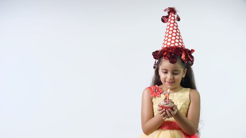 Young sweet girl from India in a beautiful birthday dress and a red party hat - birthday celebration concept. Cute Indian kid blowing a candle on a cupcake on her birthday party with a wide smile o...