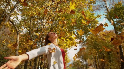SLOW MOTION, CLOSE UP, COPY SPACE: Girl stands under trees with arms outstretched as leaves come falling down from the turning treetops. Carefree Caucasian woman lets colorful tree leaves fall on her.
