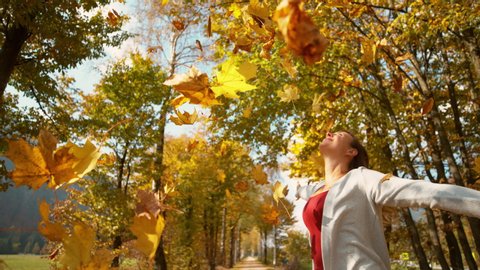SLOW MOTION, CLOSE UP, COPY SPACE: Smiling girl spins with her arms outstretched as dry tree leaves fall on her. Young Caucasian woman having fun in a picturesque autumn colored park on a sunny day.