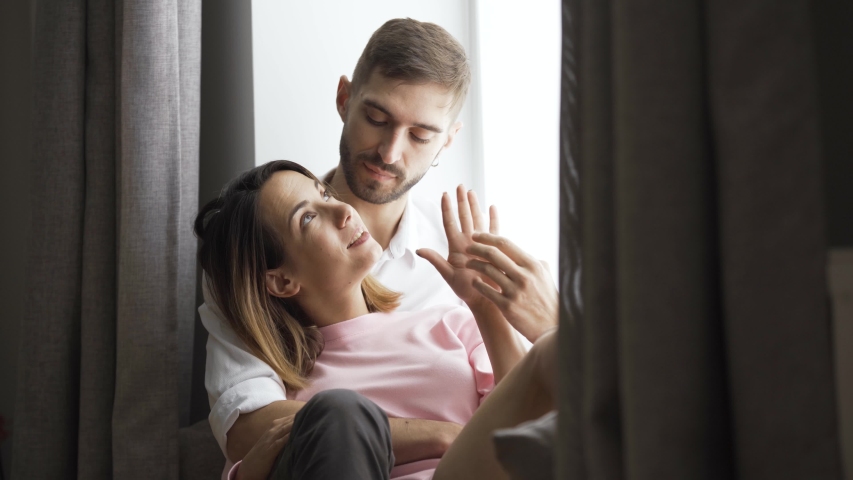 Adorable young couple is sitting by the window at home. Husband or boyfriend hugs his wife or girlfriend.