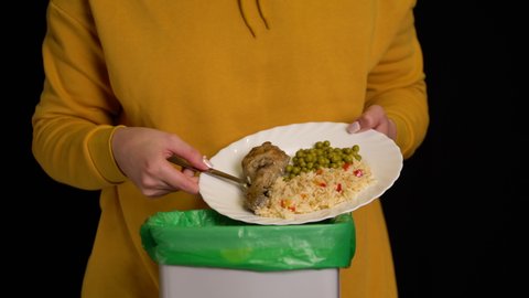 Woman scraping with a plate a chicken leg, rice, green peas into garbage bin.