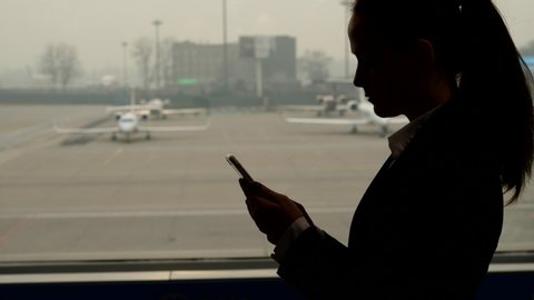 Woman stay with phone in hand, turn head and look out to apron area. Several small business aircraft parked at field of international airport. Silhouetted shot of thoughtful passenger against window