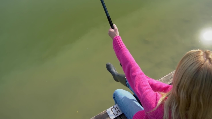 Slow motion, beautiful blonde in a pink sweater, blue jeans and rubber boots sitting on a bridge with a fishing rod by the lake in Italy. In the background is nature. Fashionable girl catches a fish