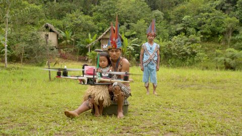 general shot of a indigenous father and his daughter flying a drone using a remote control while an old member of a tribe is watching a scene from behind in ecuador
