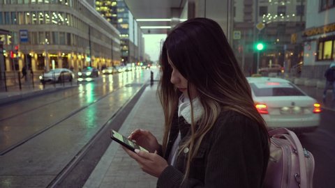 A teenager girl writes a message on her cell phone at a tram stop. In the center of Moscow, a girl in autumn rainy weather is waiting for transport. Evening movie.