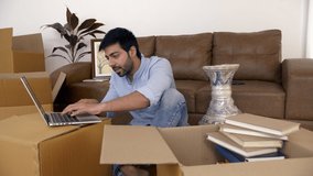 Young Indian man working on laptop amidst shifting of the new house - Relocation to metro city. Indian stock video of a young man sitting on the floor of the new house and checking emails/project r...