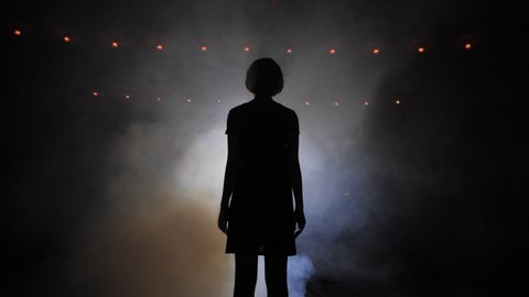 black silhouette of young woman in short dress extending hand to bright lights in smoke at darkness slow motion
