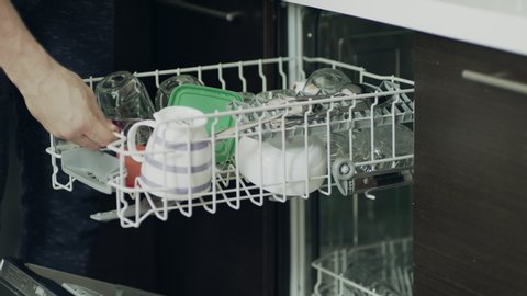 Closeup man unloading clean dishes out of dishwasher. Man hands opening dishwashing machine in slow motion. Close up male hands carrying out household chores.