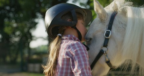 Authentic close up shot of a cute little girl with a jockey helmet is caressing and kissing a white pony horse at riding stable with a sunshine.