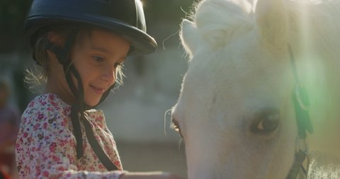 Authentic close up shot of a cute little girl with a jockey helmet is caressing a white pony horse at riding stable with a sunshine.