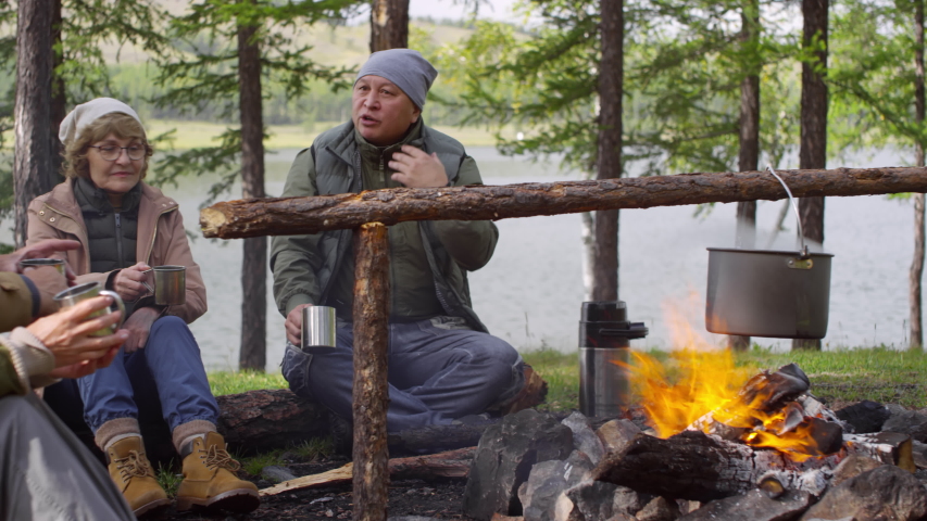 Company of senior tourists sitting by fireplace at campsite, discussing food in pot and smiling