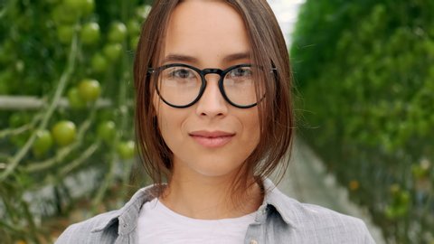 Portrait of beautiful young girl in greenhouse smiling and looking at camera. Modern farming, happy youth and profession concept. Elegant young girl in glasses.Scientist inspecting plants.
