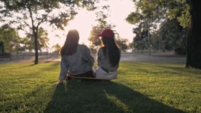 Back view of young girls friends resting and high-fiving each other while sitting on skateboard in the park