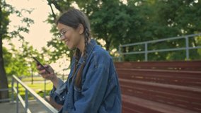 Beautiful excited young brunette girl in denim jacket smiling and chatting on smartphone while sitting in the park