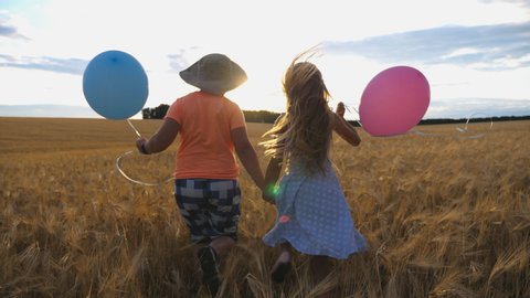 Small girl and boy holding hands of each other and running among barley plantation. Couple of little kids with balloons in arms jogging through wheat field at sunset. Concept of child love. Rear view