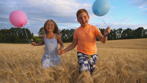Couple of happy little kids with balloons in arms jogging through wheat field. Small girl and boy holding hands of each other and running among barley plantation. Concept of child love. Slow motion