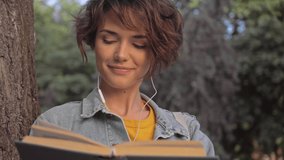 Attractive pleased young brunette woman in denim jacket listening music with earphones and reading book while leaning on a tree in the park