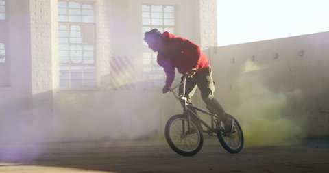 Side view of two young Caucasian men riding and doing tricks on BMX bikes with purple and yellow smoke grenades attached to them, in an abandoned warehouse