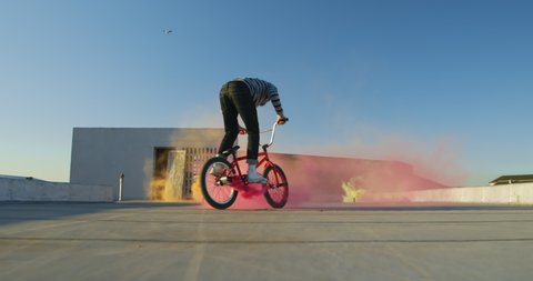 Side view of a young Caucasian man riding a BMX bike and doing tricks on the rooftop of an abandoned warehouse, with a pink smoke grenade attached to the bike  in slow motion