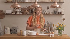 Cheerful young blonde woman with long curly hair listening music with earphones on smartphone and dancing while cooking at the kitchen
