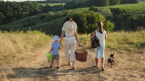 Family time in nature. Mother and her daughter and little son holding hands going to a picnic with a wicker basket, plaid, and dog. Rear view, slow motion shot