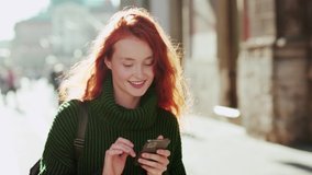 Beautiful young woman with wavy red hair browsing in her phone smiling and walking in the city streets looking around positive emotion - Powered by Shutterstock - Get 15% off with code: PIKWIZARD15