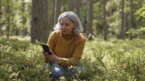 Elderly Caucasian woman wearing jeans and sweater sitting on her haunches in woods and making photos using telephone